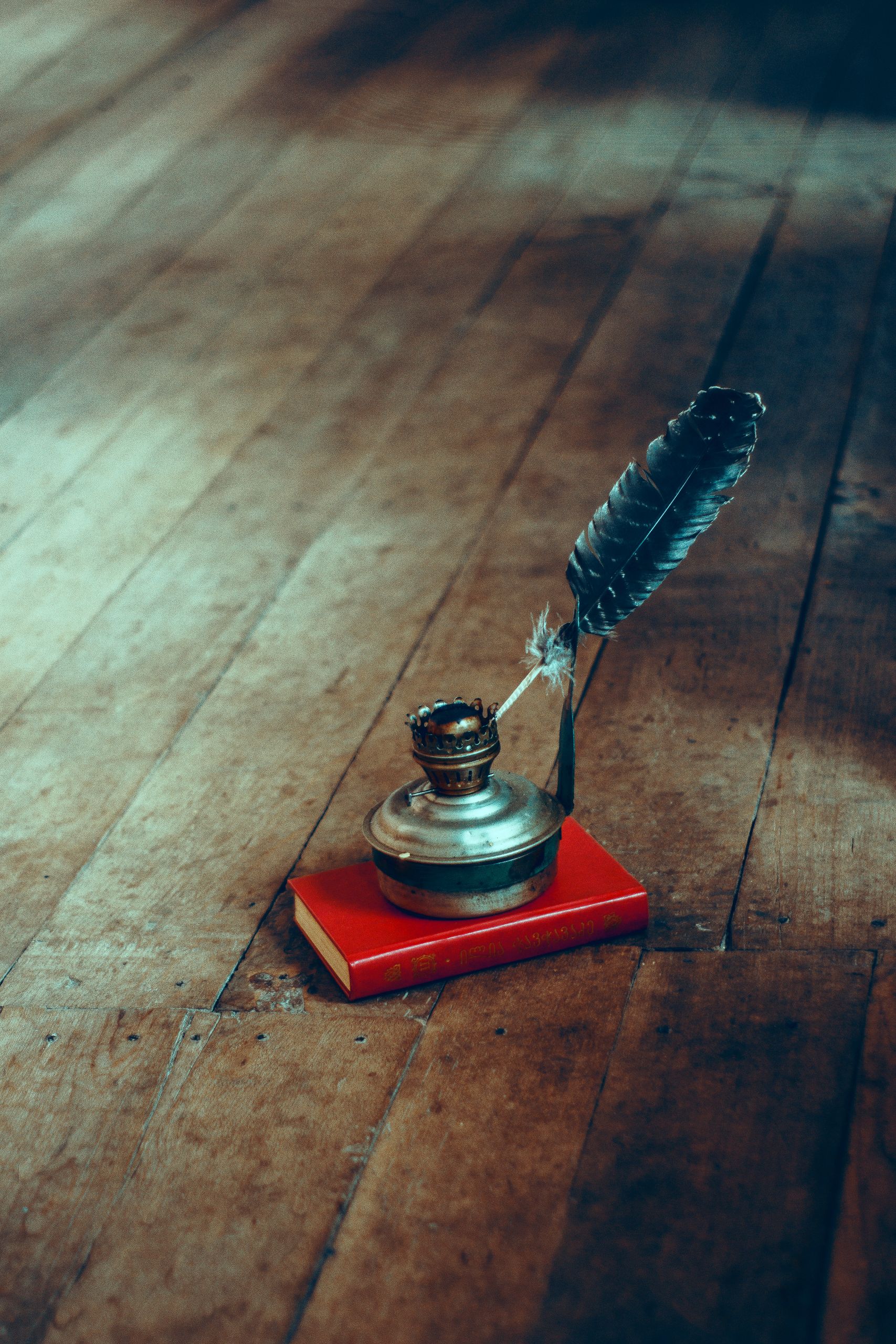 An antique ink pot with a feather quill resting on a red book on wooden floorboards.